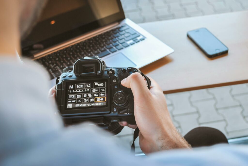 Mensch bedient Spiegelreflexkamera dabei schaut man über seine Schulter im Hintergrund ist ein Laptop und ein Handy