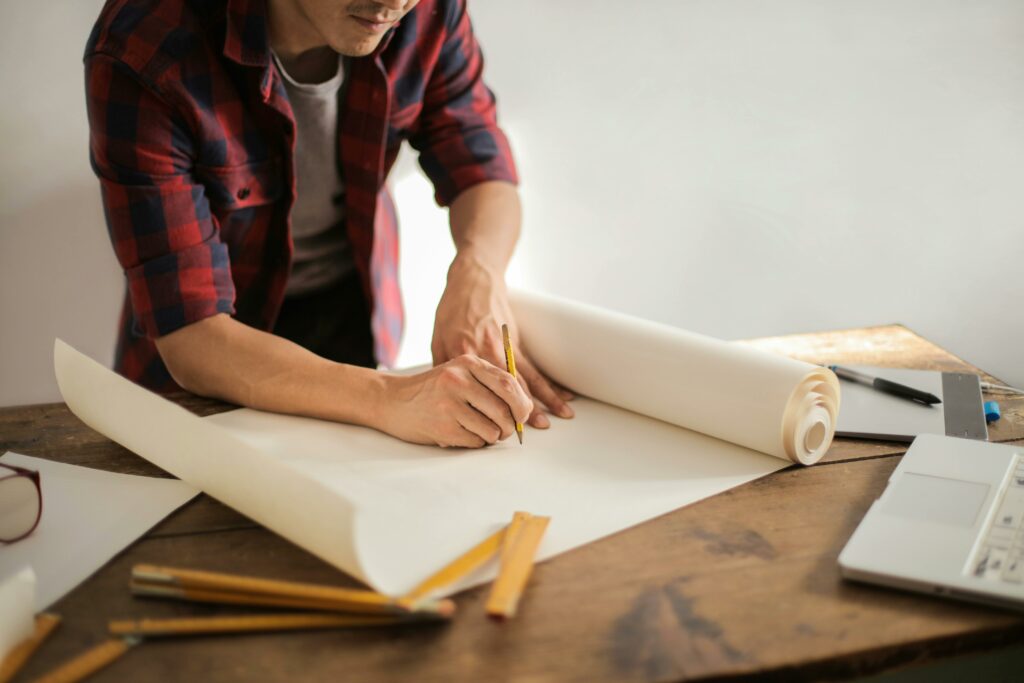 Mann zeichnet auf einer großen Rolle Papier, um ihn herum liegen Lineale und ein Maßstab und Büromaterial.